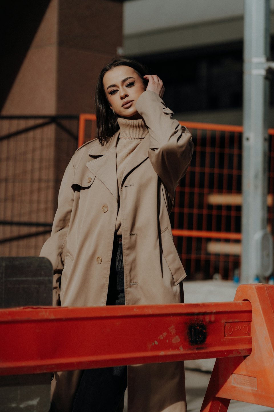 Woman in a beige trench coat stands near urban buildings.
